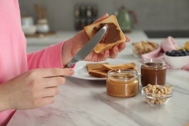 Woman spreading tasty nut butter onto toast at white marble table, closeup