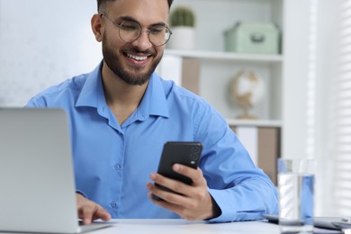 Photo of Happy young man using smartphone while working with laptop at white table in office