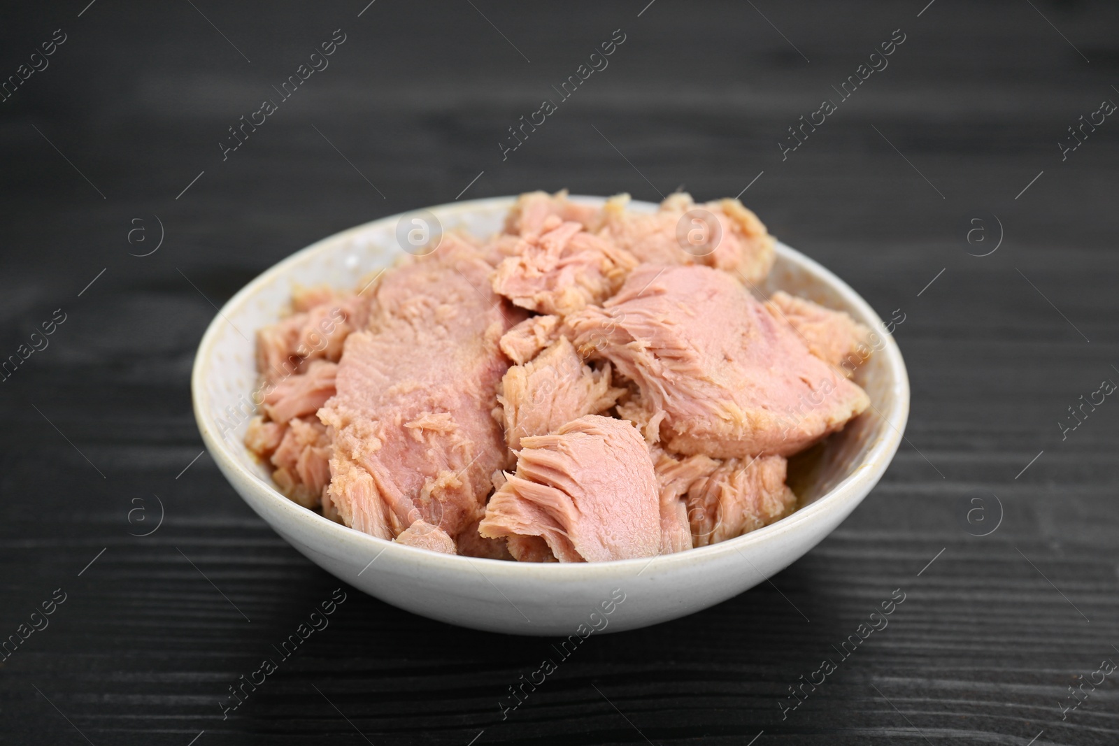 Photo of Bowl with canned tuna on black wooden table, closeup