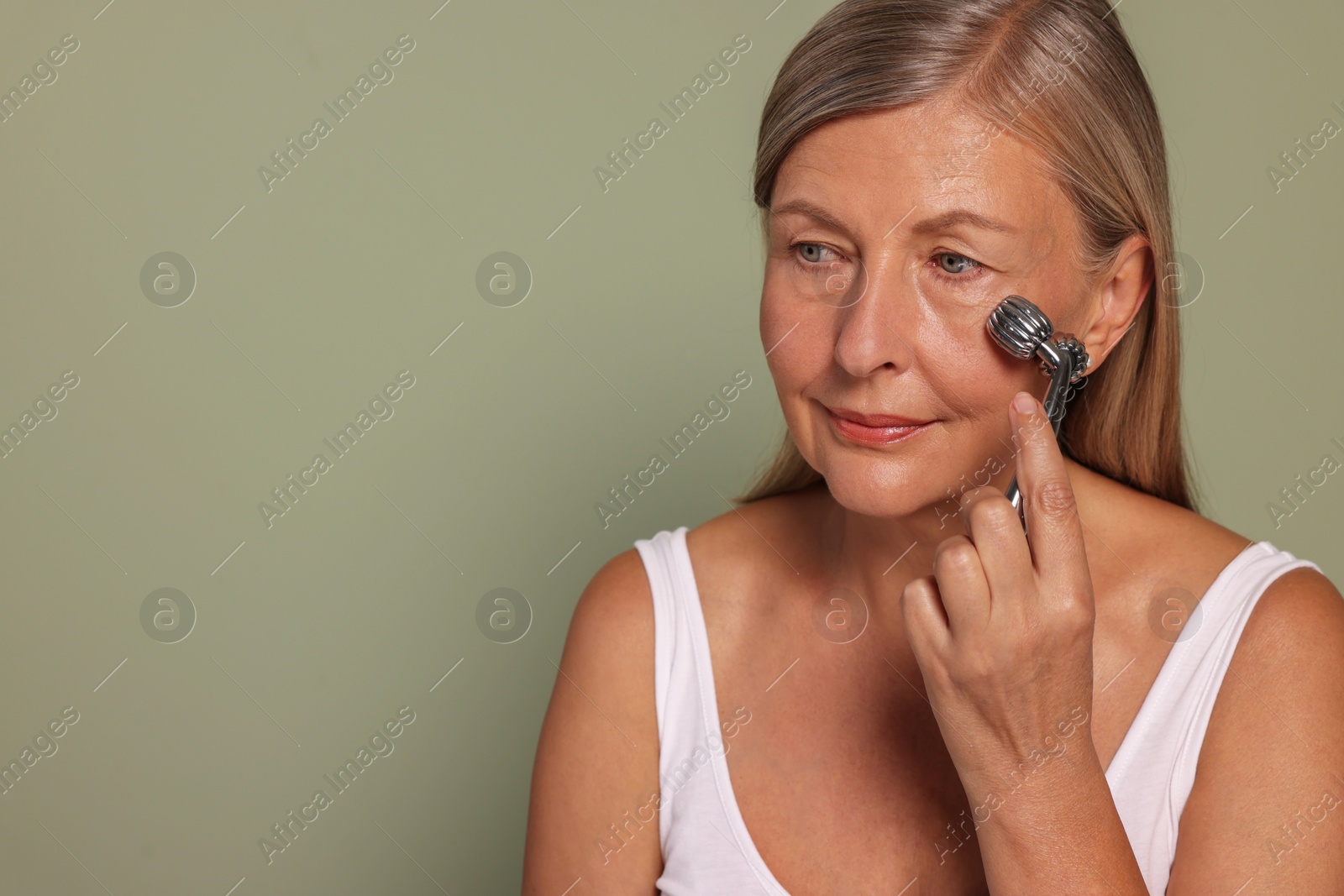 Photo of Woman massaging her face with metal roller on green background. Space for text