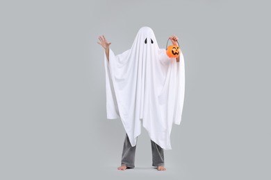 Photo of Woman in white ghost costume holding pumpkin bucket on light grey background. Halloween celebration