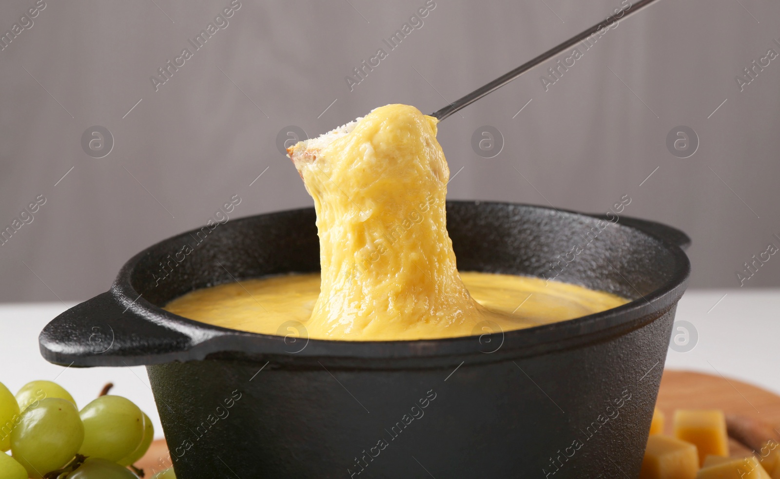 Photo of Dipping piece of bread into fondue pot with tasty melted cheese at table against gray background, closeup