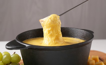 Dipping piece of bread into fondue pot with tasty melted cheese at table against gray background, closeup