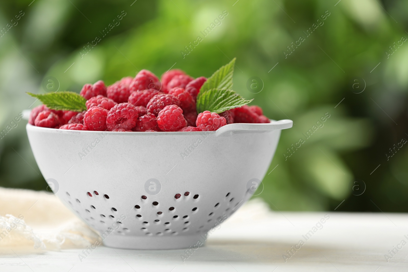 Photo of Colander with delicious ripe raspberries on white table against blurred background, space for text