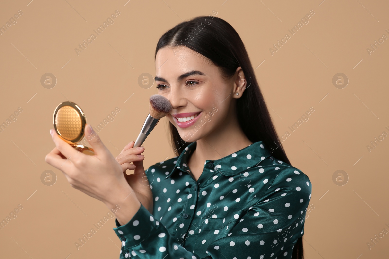 Photo of Happy woman with cosmetic pocket mirror applying makeup on light brown background
