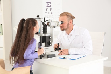 Photo of Children's doctor examining little girl with ophthalmic equipment in clinic