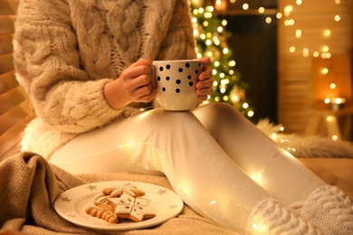 Photo of Woman with cup of hot drink and cookies near Christmas tree at home, closeup