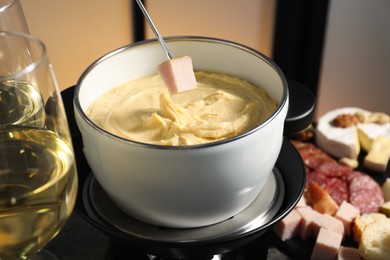 Dipping piece of ham into fondue pot with melted cheese at table with snacks, closeup