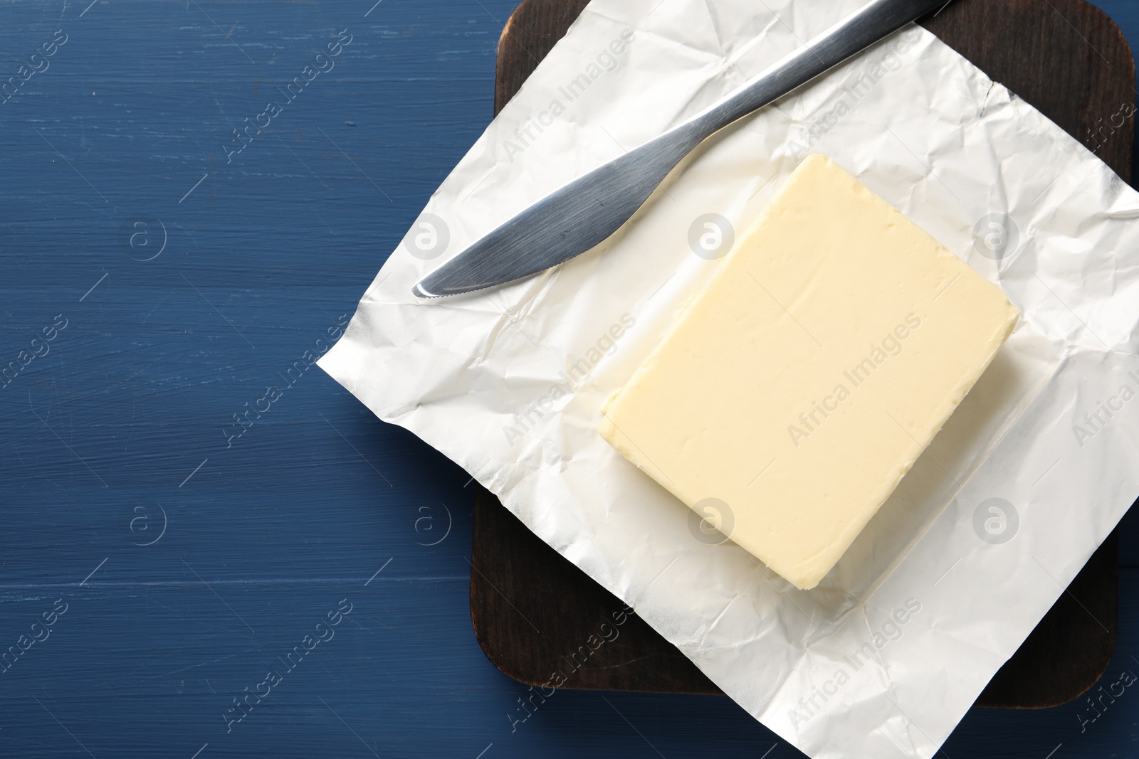 Photo of Block of tasty butter in open foil packaging and knife on blue wooden table, top view