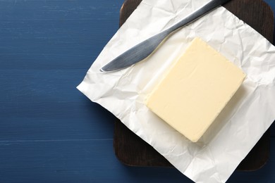 Block of tasty butter in open foil packaging and knife on blue wooden table, top view