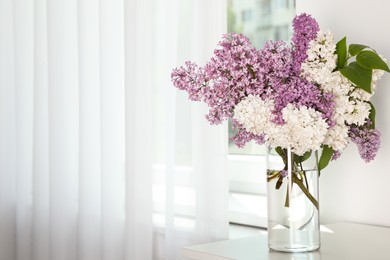 Photo of Beautiful lilac flowers in glass vase on white table near window. Space for text