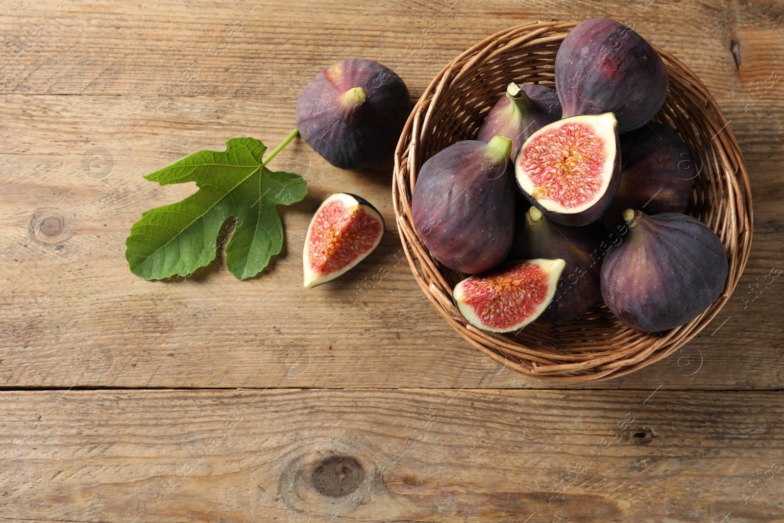 Photo of Wicker bowl with fresh ripe figs and green leaf on wooden table, flat lay. Space for text