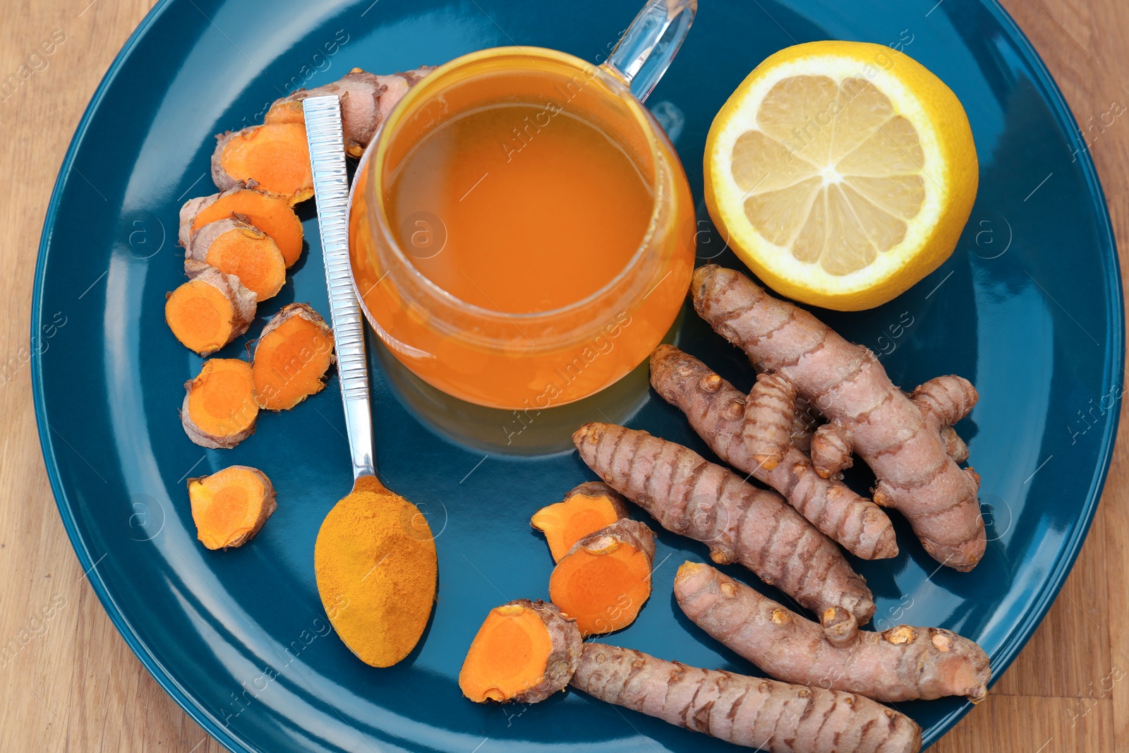 Photo of Plate with glass cup of hot tea, lemon, turmeric powder and roots on wooden table, above view