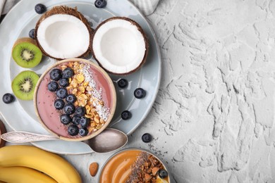 Bowl of delicious fruit smoothie with fresh blueberries, granola and different ingredients on white textured table, flat lay. Space for text