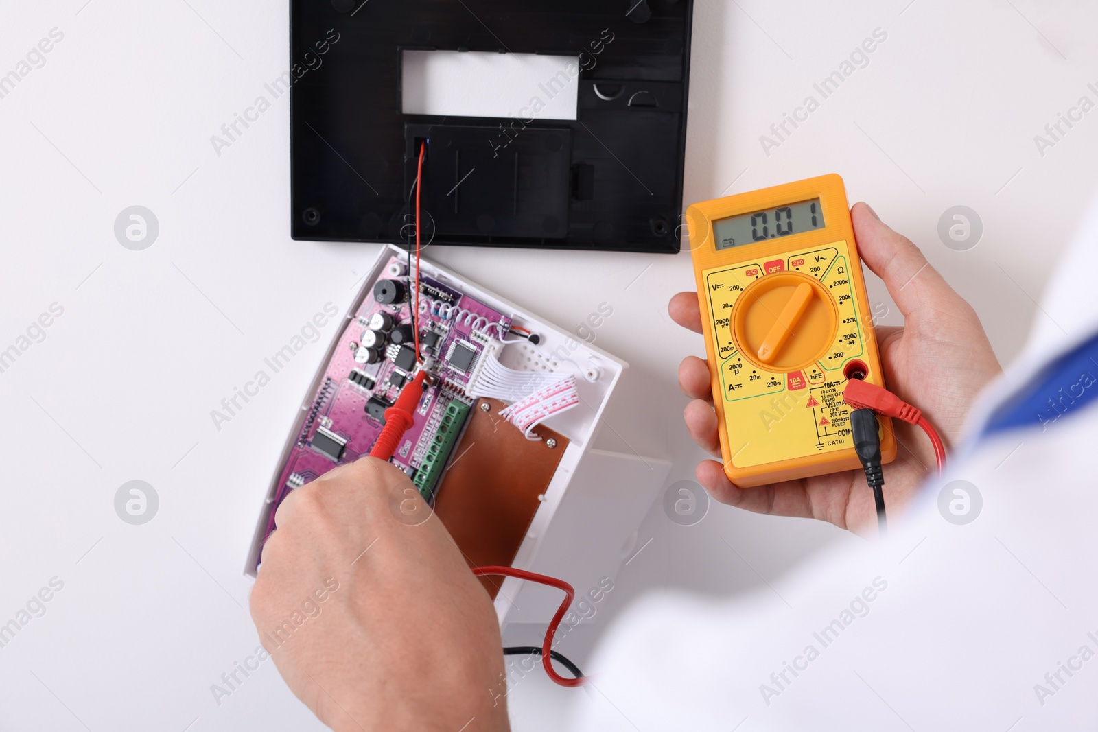 Photo of Man using digital multimeter while installing home security alarm system on white wall indoors, closeup