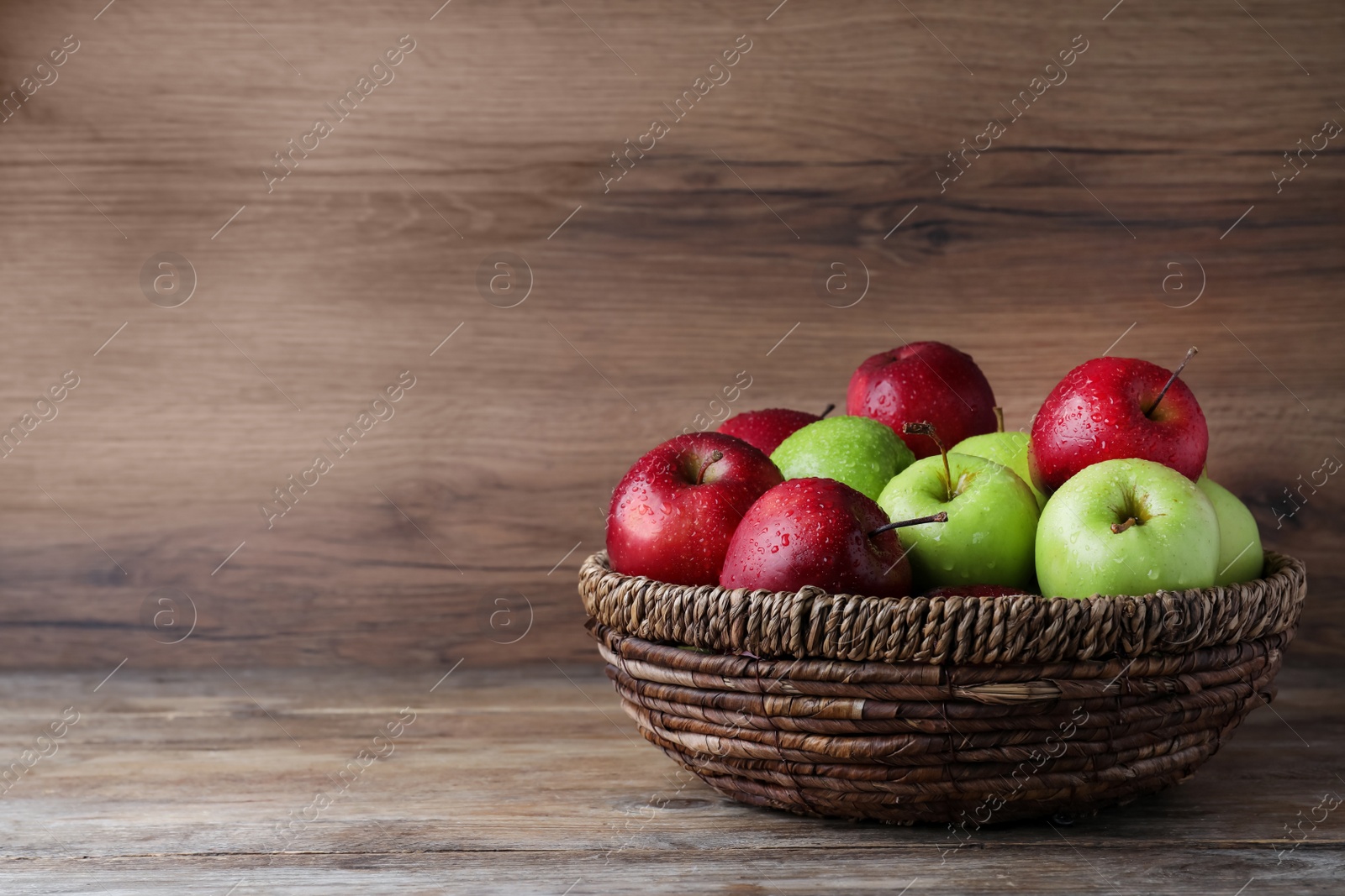 Photo of Fresh ripe green and red apples with water drops in wicker bowl on wooden table, space for text