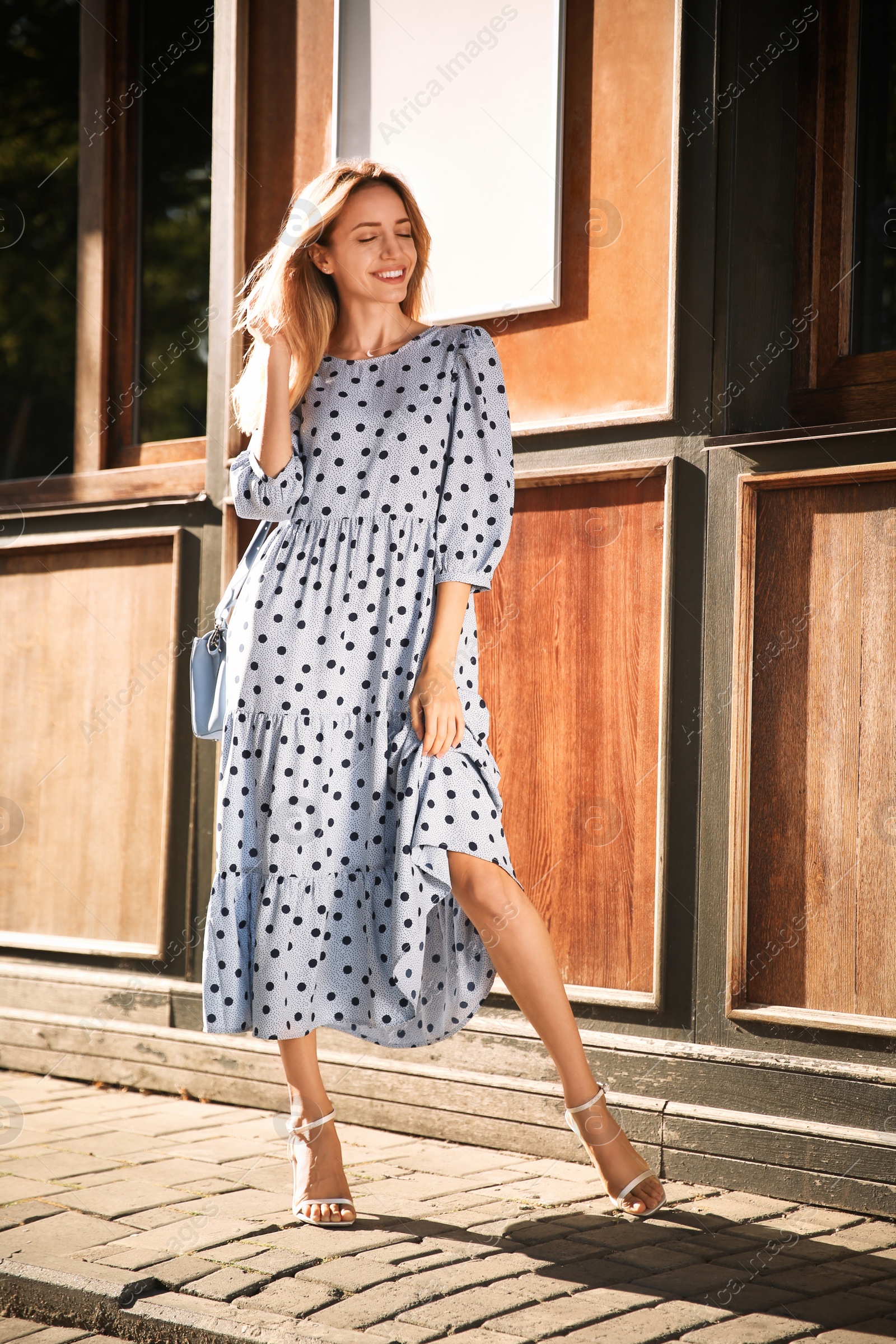 Photo of Beautiful young woman in stylish light blue polka dot dress with handbag on city street