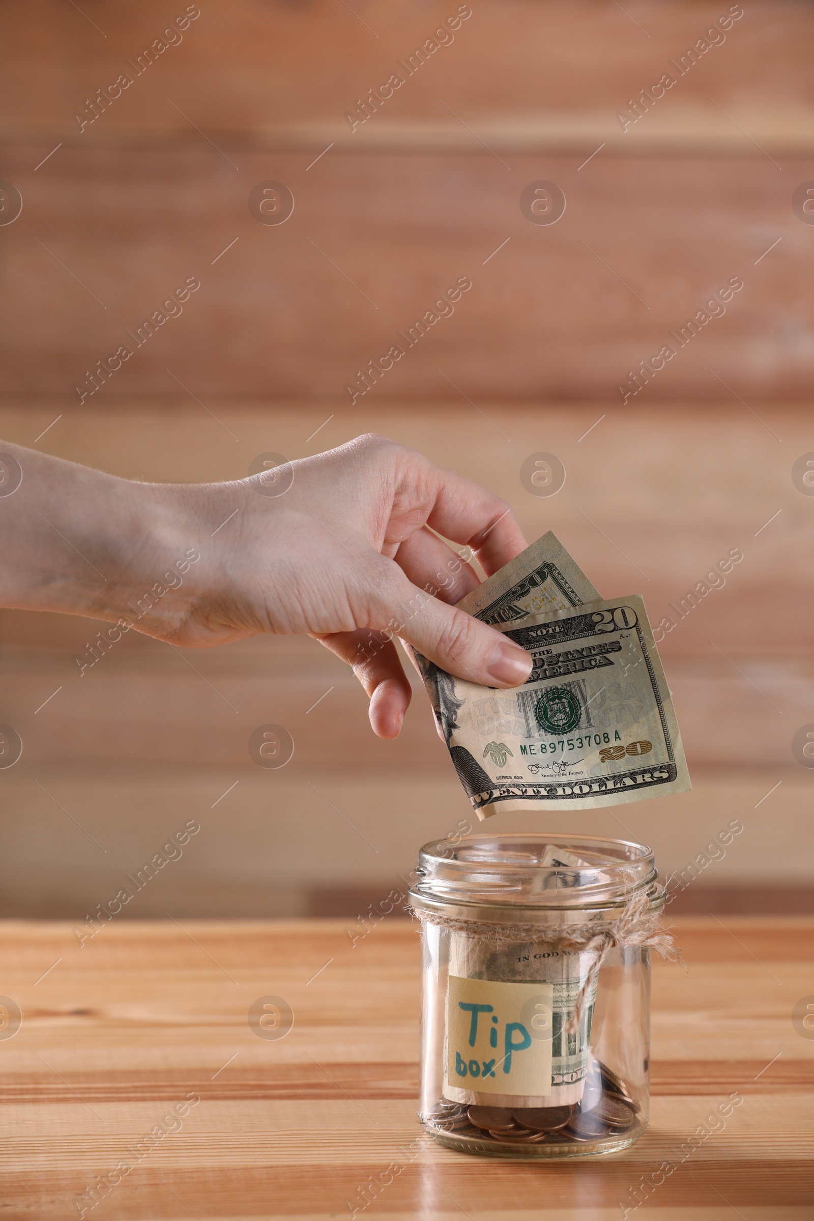 Photo of Woman putting tips into glass jar on wooden table, closeup