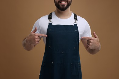 Photo of Smiling man pointing at kitchen apron on brown background, closeup. Mockup for design