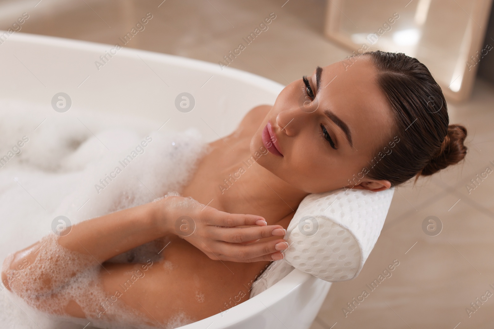 Photo of Young woman using pillow while enjoying bubble bath indoors