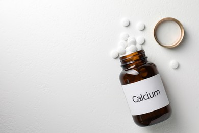 Photo of Overturned bottle of calcium supplement pills on white table, flat lay. Space for text