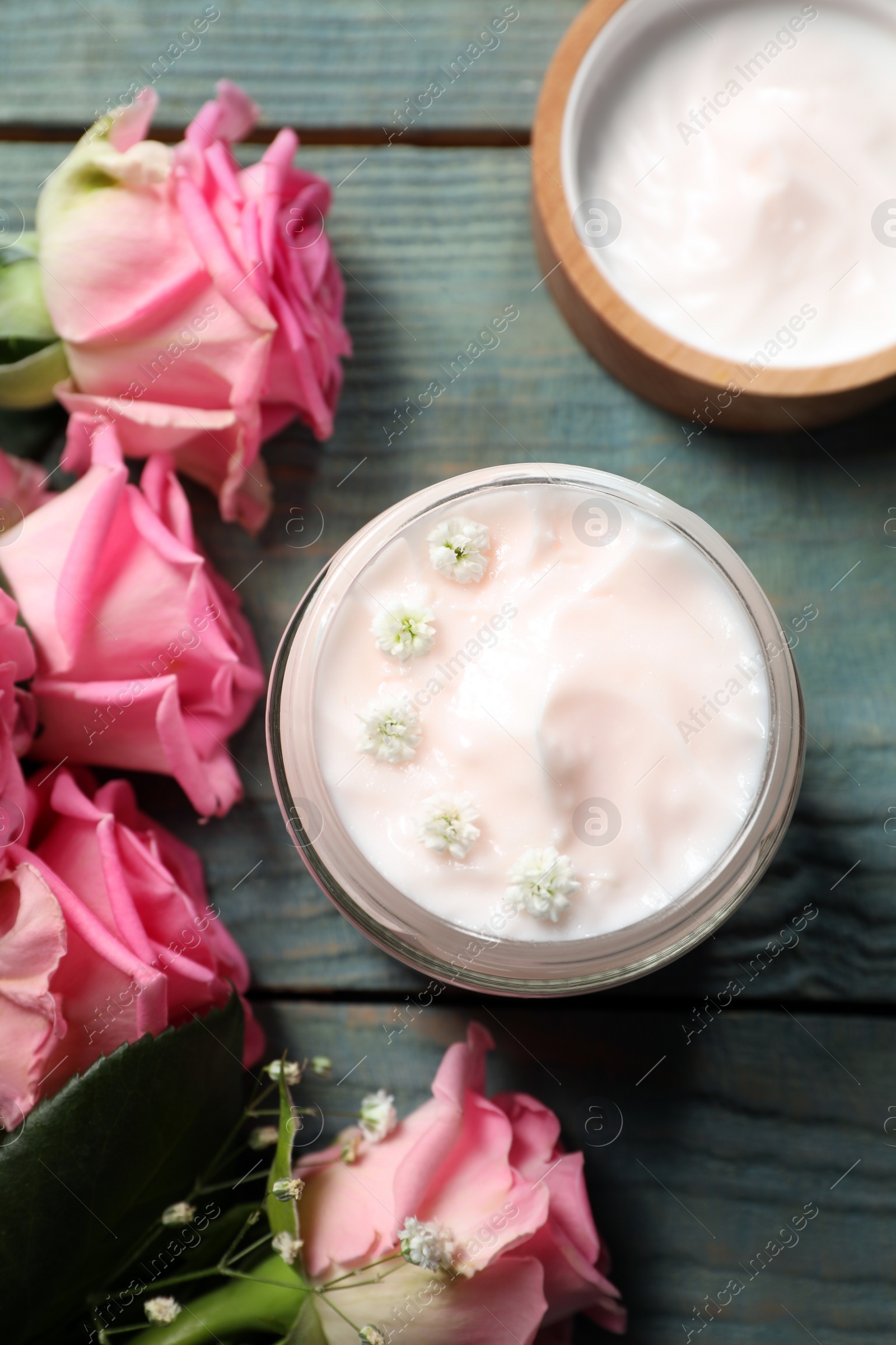 Photo of Jar of face cream and pink roses on light blue wooden table, flat lay