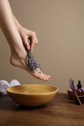Photo of Woman with lavender flowers holding her foot over bowl of water on wooden floor, closeup. Pedicure procedure