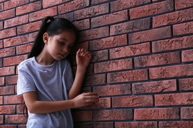 Photo of Sad little girl near brick wall, space for text. Child in danger