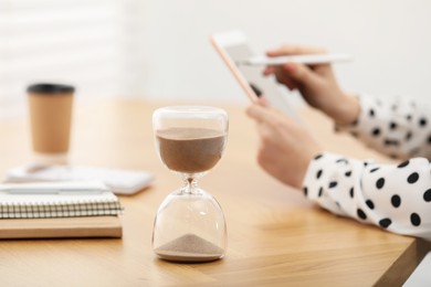 Photo of Hourglass with flowing sand on desk. Woman using tablet indoors, selective focus