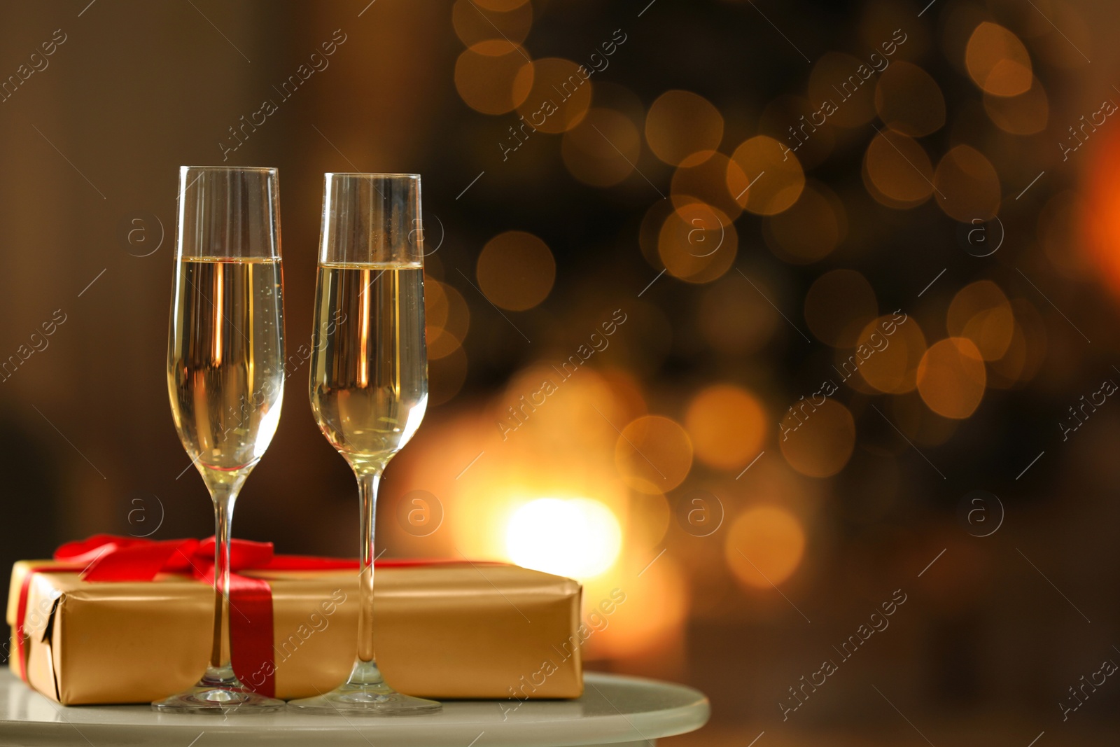 Image of Glasses with champagne and Christmas gift on table in room, bokeh effect. Space for text