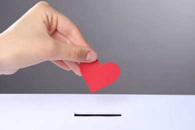 Photo of Woman putting red heart into slot of donation box against grey background, closeup