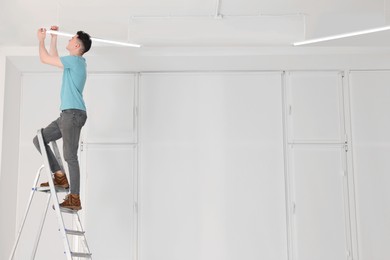 Young man installing ceiling lamp on stepladder indoors