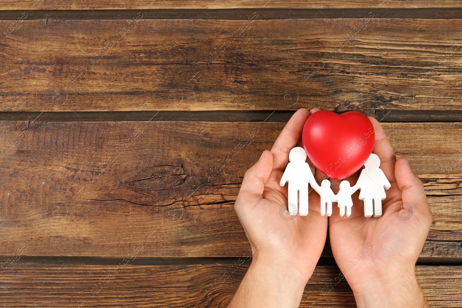 Photo of Young man holding family figure and red heart in his hands on wooden background, top view. Space for text