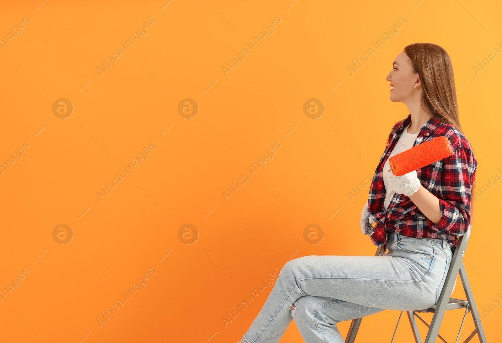 Photo of Designer with roller sitting on folding ladder near freshly painted orange wall, space for text