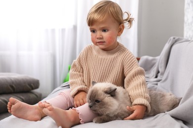 Image of Cute little child with allergic redness sitting with adorable pet on sofa at home