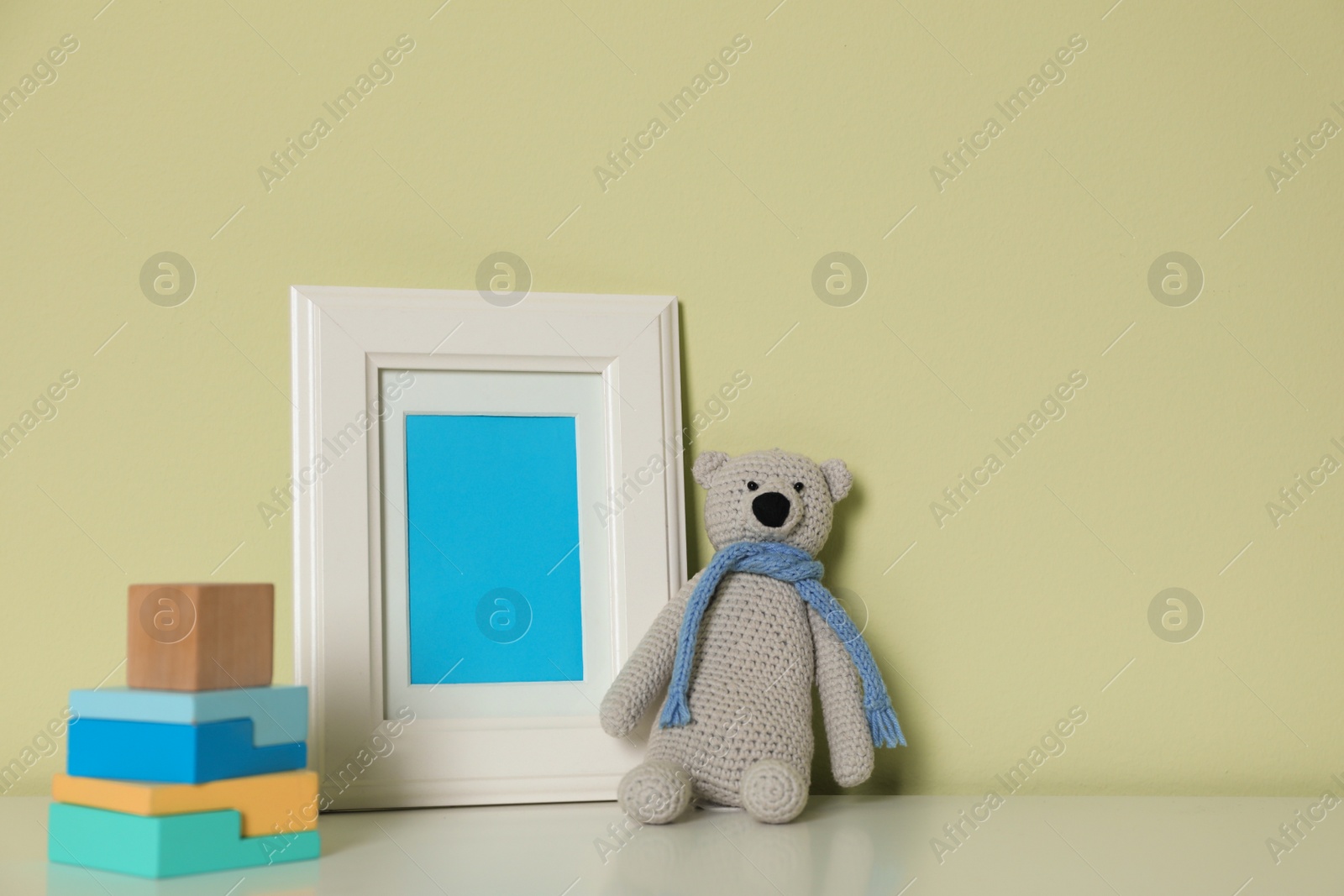 Photo of Empty photo frame, toy bear and building blocks on white table near light green wall. Space for design