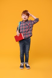 Confused schoolboy with backpack and book on orange background