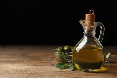 Photo of Glass jug of oil, ripe olives and green leaves on wooden table. Space for text