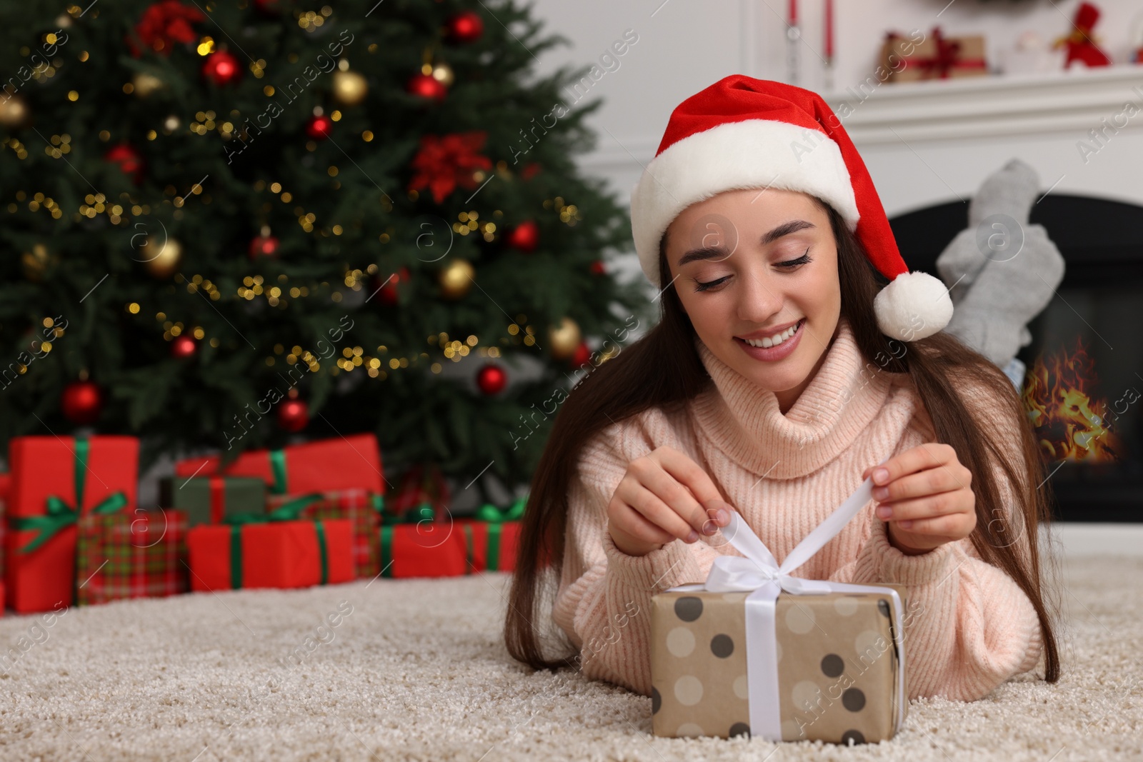 Photo of Happy young woman in Santa hat opening gift box in room decorated for Christmas