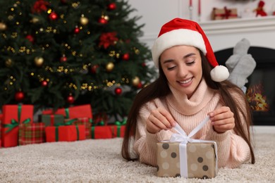 Photo of Happy young woman in Santa hat opening gift box in room decorated for Christmas