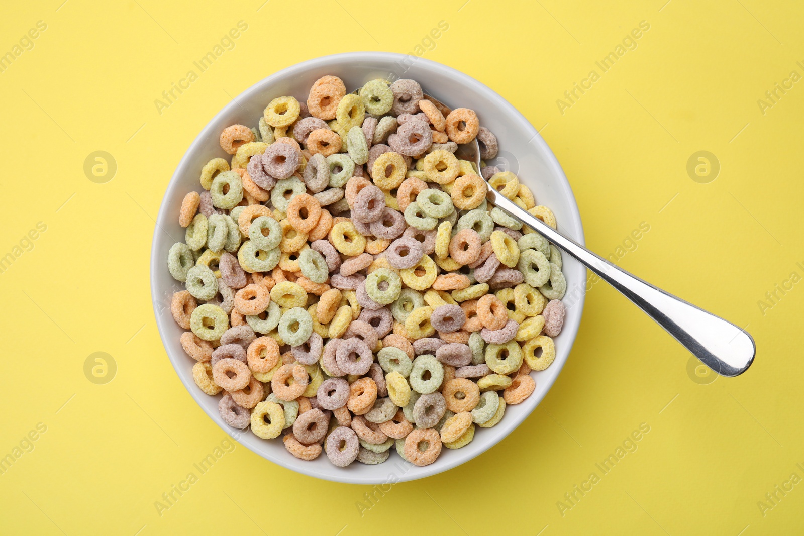 Photo of Tasty cereal rings in bowl and spoon on yellow table, top view