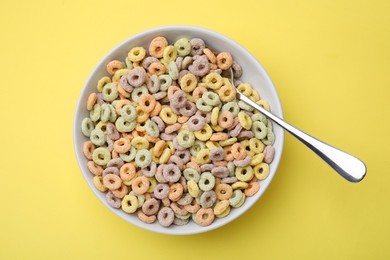 Photo of Tasty cereal rings in bowl and spoon on yellow table, top view