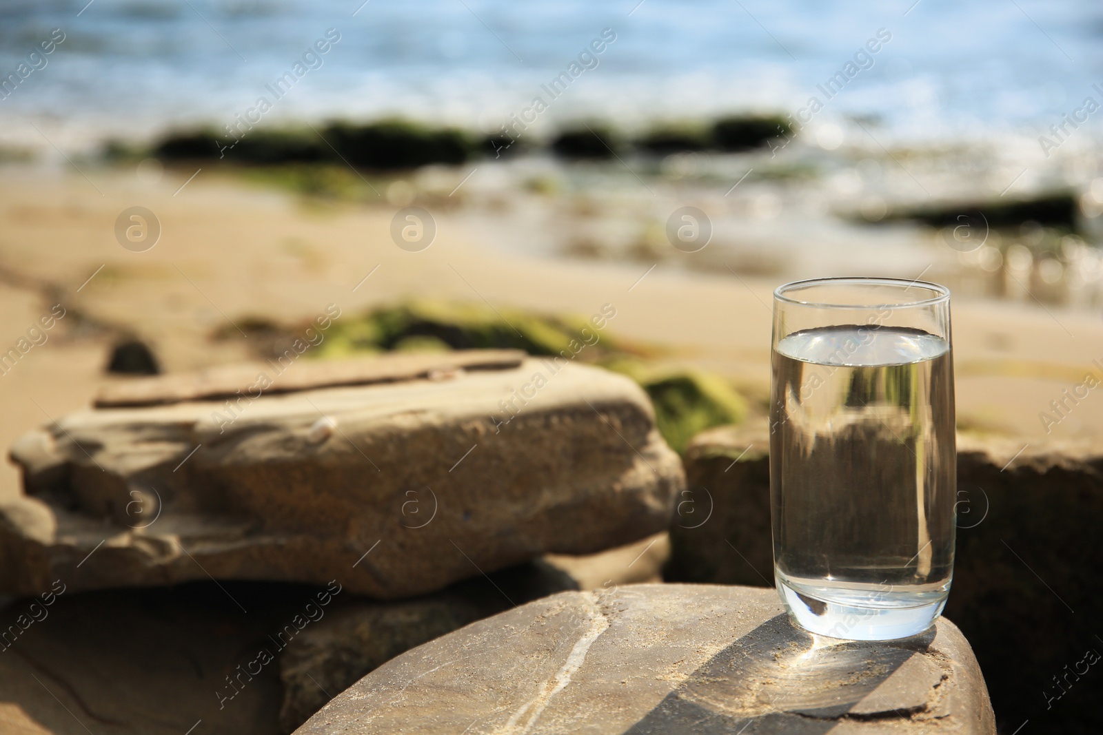 Photo of Glass of fresh water on stone near sea, space for text