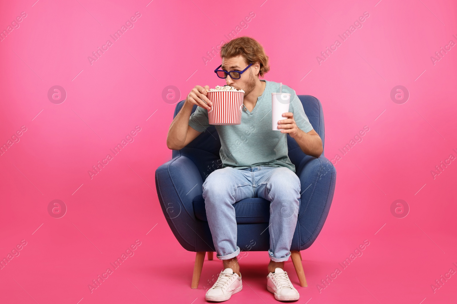 Photo of Man with 3D glasses, popcorn and beverage sitting in armchair during cinema show on color background