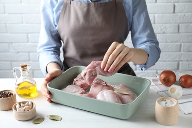 Photo of Woman putting raw rabbit meat into baking dish at white wooden table, closeup