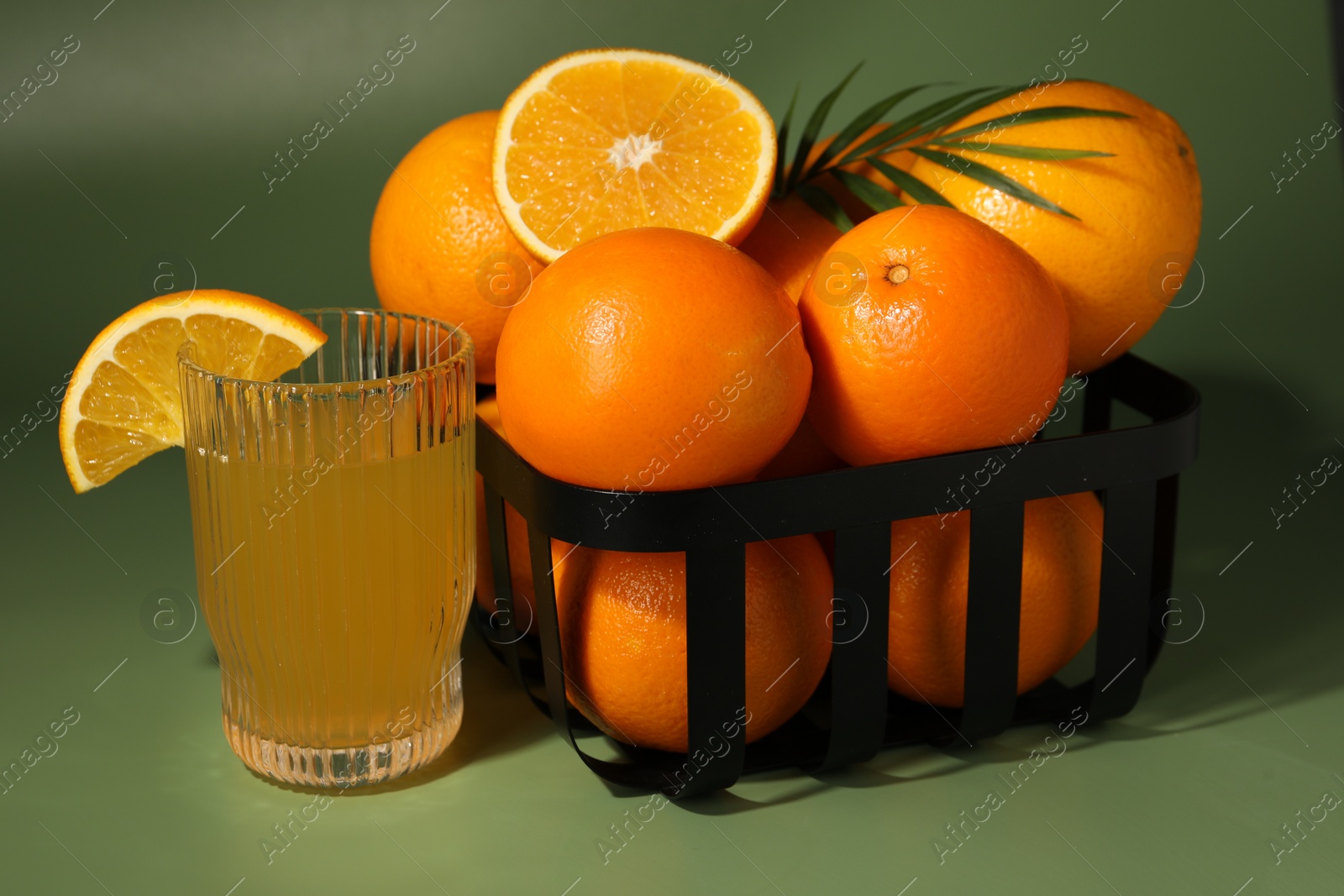 Photo of Fresh oranges in metal basket and glass of juice on green background, closeup