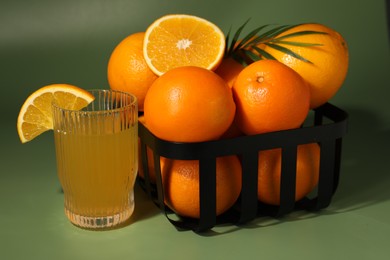 Fresh oranges in metal basket and glass of juice on green background, closeup