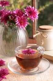 Photo of Beautiful chrysanthemum flowers and cup of tea on beige textured table near window. Autumn still life