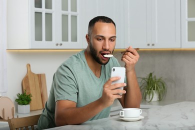 Photo of Young man using smartphone while having breakfast at white marble table in kitchen. Internet addiction