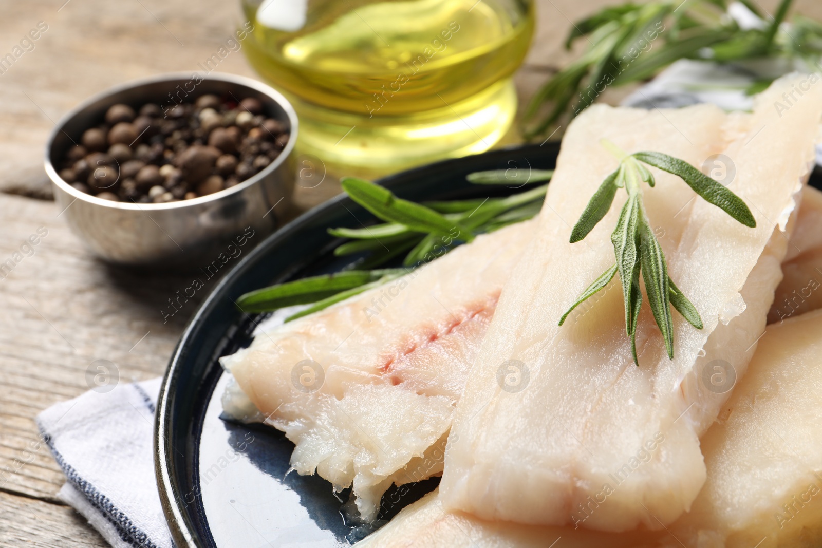 Photo of Pieces of raw cod fish, rosemary and peppercorns on wooden table, closeup